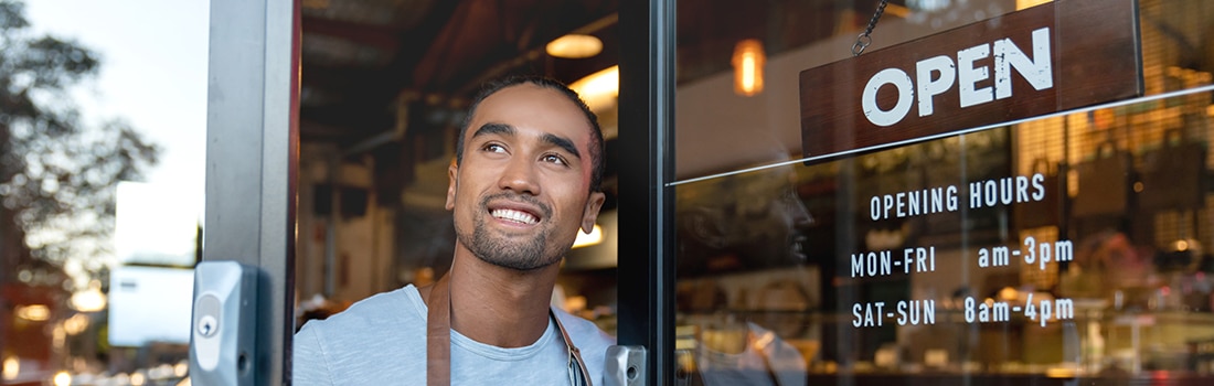 a guy smiling while opening the door of a business with a sign that says ‘open'